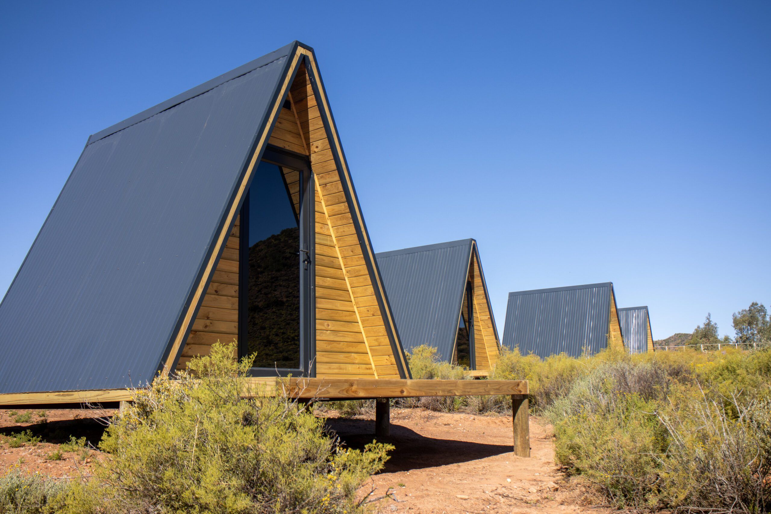 A row of timber cabins with steep triangular gables and dark metal roofs, standing in low bush under a deep blue sky