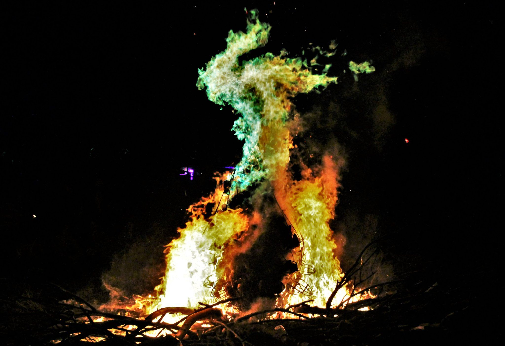 A tall bonfire burning against a black night sky, embers rising from the flames