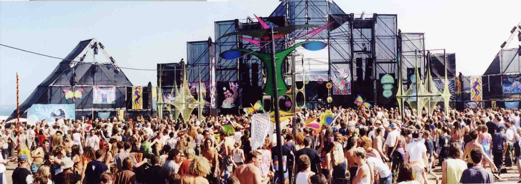 Panoramic archive frame from Silverstroom 1996: the crowd dancing in daylight before scaffold stage towers hung with psychedelic banners