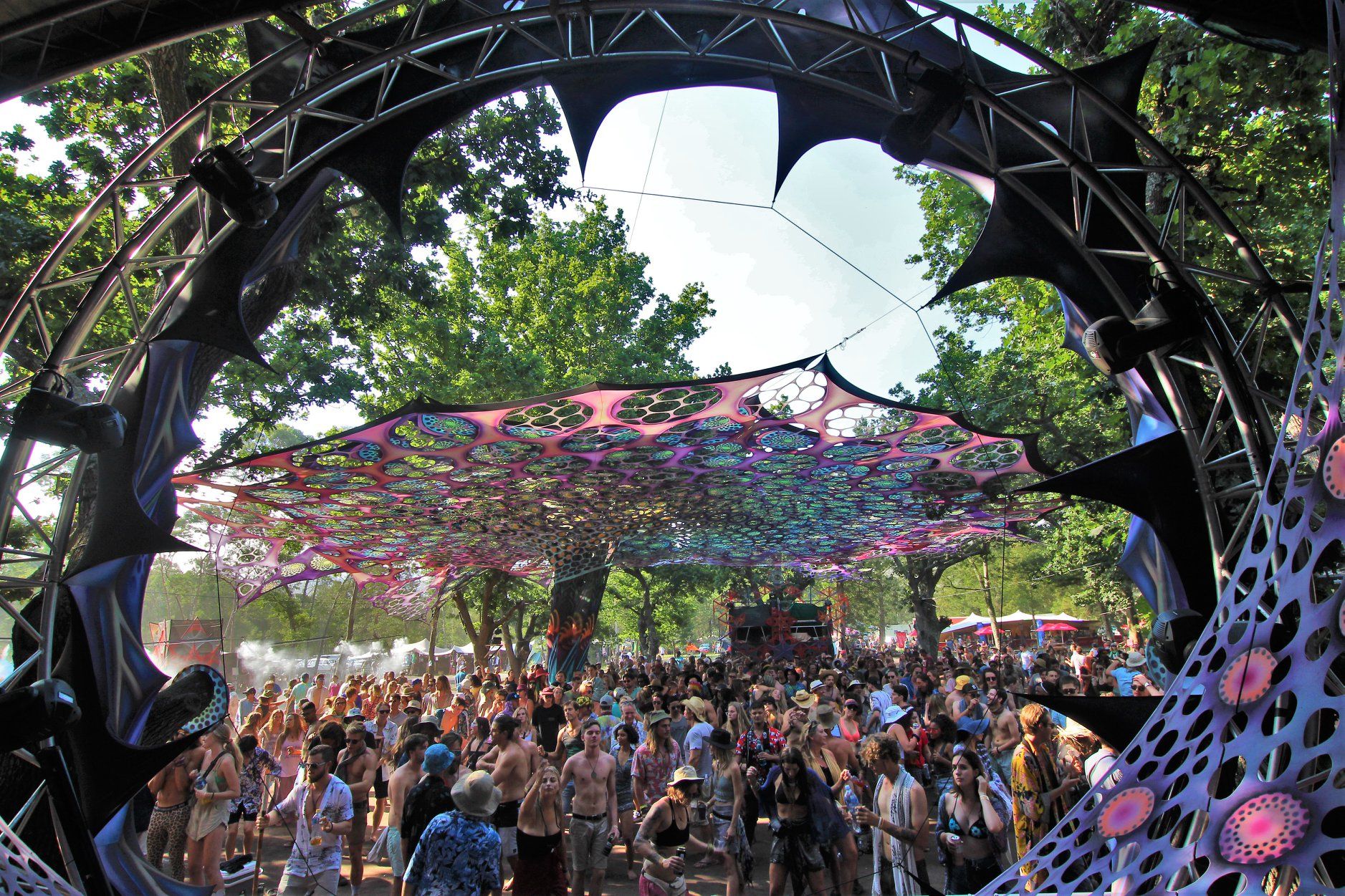 Daytime dancefloor crowd beneath a psychedelic canopy, framed by the stage arch and summer trees