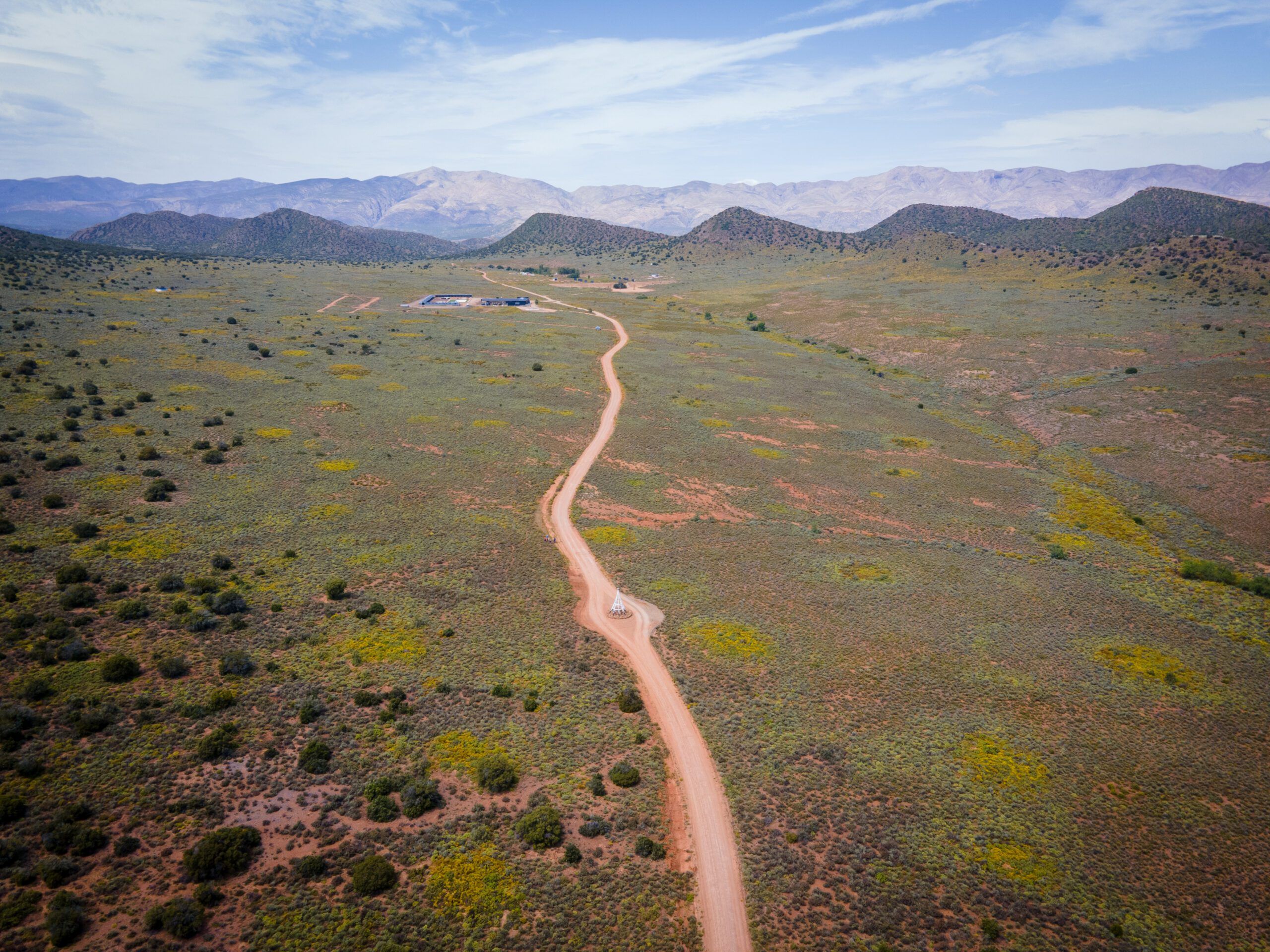 An aerial view of a dirt road curving through green Karoo veld towards a long line of mountains under a wide, pale sky