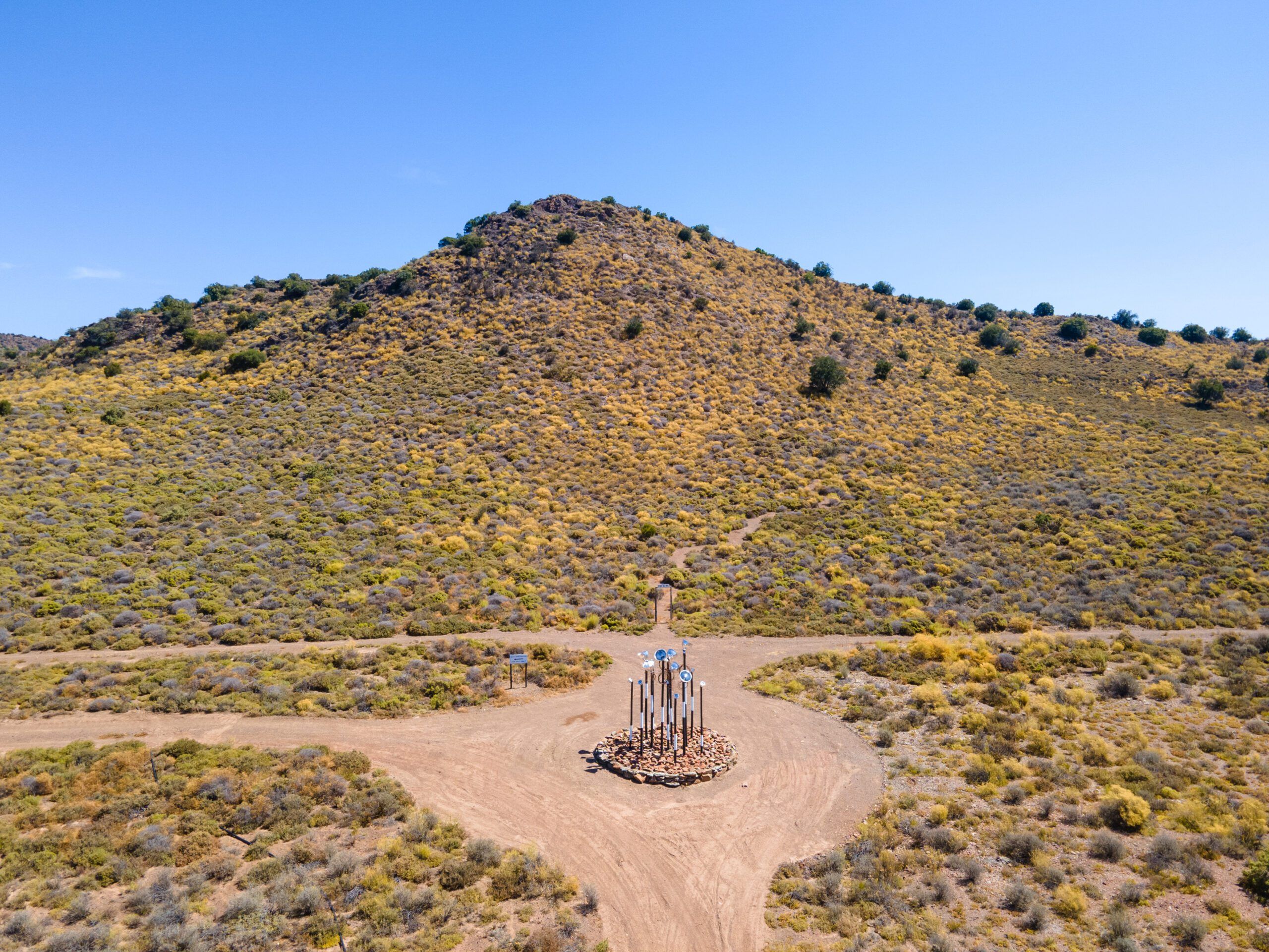 A flowering hillside above a dirt track junction, with a small sculpture of mirrored discs set on a circle of stones