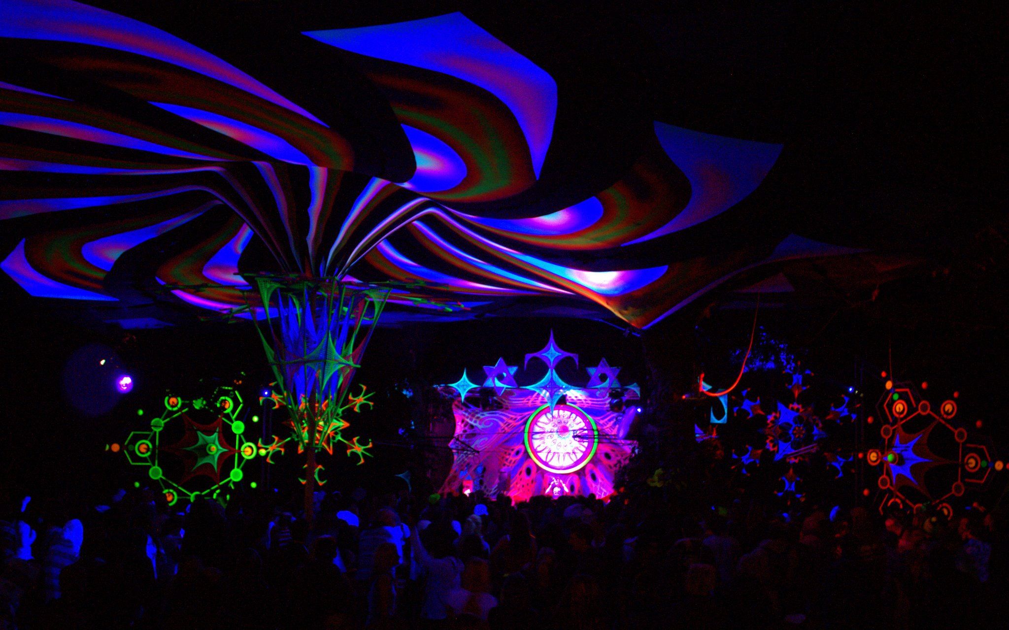 Night dancefloor beneath swirling UV-lit canopy sails, string-art sculptures glowing and a mandala stage at the far end