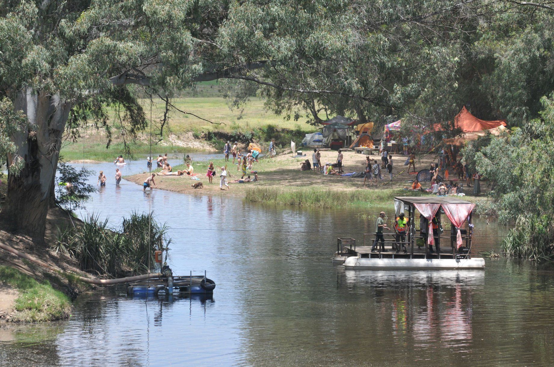 Festival-goers swimming and resting on a shaded riverbank beside the camp, a small raft crossing the water
